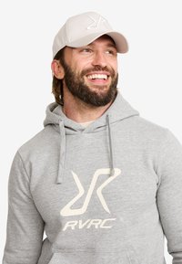 Grå sweatshirt med huva, med en stor krämfärgad logotyp "RVRC" i mitten. En ljusgrå basebollkeps fulländar outfiten.