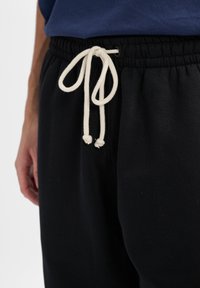 Schwarze Jogginghose mit elastischem Bund und weißem Zugband. Das Material wirkt weich und strukturiert, mit einem lässigen Schnitt.
