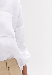 Camicia bianca in lino con taschino sul petto e maniche arrotolate, abbinata a pantaloni beige con un bottone decorativo in vita.