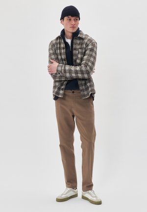 Geruite button-up overhemd in beige en bruin, marineblauwe zip-up trui, tan chino's, witte sneakers met groene accenten en een marineblauwe beanie.