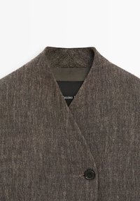 Blazer marron texturé avec un col sans cran et une fermeture à un bouton noir, laissant entrevoir une partie de la doublure intérieure noire.