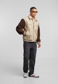 Jachetă varsity bej și maro cu guler, nasturi tip snap și manșete stripe. Asortată cu blugi închiși la culoare și teniși multicolori.