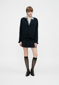 Černý pletený svetr s výstřihem do V, přes šedou košili s límečkem. V kombinaci s černou sklady sukni a průhlednými černými punčochami.