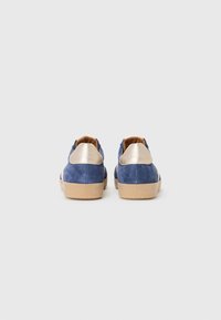 Blå mockasinsneakers med en gylden metallisk hælinddækning, struktureret overflade og en lys brun gummisål. Bagfra vist.