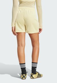 Lichtgele sportshorts met een elastische tailleband, gecombineerd met zwarte en witte gestreepte sokken en gouden schoenen, zichtbaar vanuit de achterkant.