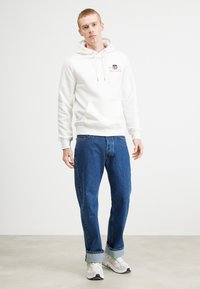 Haut à capuche crème avec une poche avant et des cordons de serrage, associé à un jean bleu et des baskets blanches. Style simple et décontracté avec une coupe ample.
