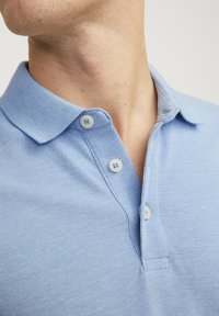 Hellblaues Polohemd aus strukturiertem Stoff, mit einer Knopfleiste mit zwei Knöpfen und einem Kragen. Verfügt über weiße Knöpfe und einen leicht taillierten Schnitt.