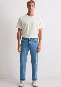 Mężczyzna stojący przy białej ścianie, ubrany w gładką, kremową koszulkę, jasnoniebieskie dżinsy i białe trampki z beżowymi akcentami.