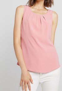 Blusa sin mangas de color rosa claro con detalle plisado en el escote, hecha de una tela suave, con dobladillo redondeado y un corte relajado.