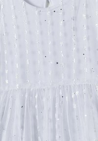 Robe blanche avec une superposition en dentelle texturée, ornée de sequins argentés et de petits points noirs, avec une jupe en tulle froncée au bas.