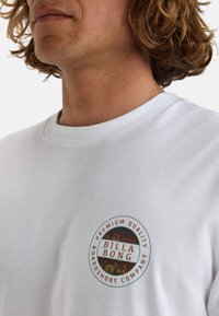 Biała koszulka z krótkim rękawem z okrągłym logo z napisem "BILLA BONG" w czerwonym kolorze i "PREMIUM QUALITY BOARDSHORT COMPANY" w czarnym.