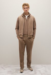 Grauer Trainingsanzug mit Reißverschlussjacke und sportlichen Hosen. Weiches Material, lockere Passform, mit gerippten Bündchen und hohem Kragen. Sneaker-Details.