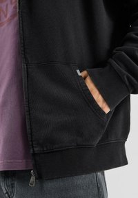 Czarna bluza z kapturem zapinana na zamek, z prążkowymi ściągaczami na rękawach i u dołu, z boczną kieszenią na zamek oraz kieszenią z przodu, noszona na fioletowej koszulce.