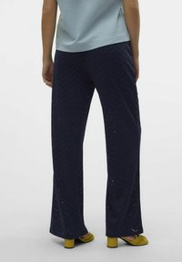 Tummansininen kuvioitu housut, joissa on siksak-tekstuuri ja vetoleikkaukset, yhdistettynä keltaisiin paksuheelisiin kenkiin.