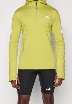 Langärmliges gelbes Kapuzen-Shirt mit Viertelreißverschluss und Daumenlöchern, kombiniert mit schwarzen Sportshorts, beide mit weißem Adidas-Logo.
