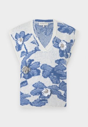 Femme portant une chemise blanche avec une broderie florale bleue, un gilet en maille bleu et blanc à motifs floraux, un jean large bleu clair et des chaussures noires à bout pointu.