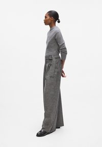 Grauer Pullover mit Rundhalsausschnitt, kombiniert mit weit geschnittenen grauen Hosen, die mit einem Gürtel und Knopfdetails versehen sind. Die Schuhe sind schwarz mit Nietenakzenten.