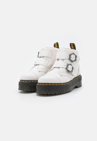 Bottines blanches en cuir avec deux sangles Velcro, présentant un matériau texturé, une semelle noire épaisse et des accents de couture jaune.