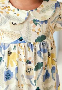 Niña vistiendo un vestido crema con un collar tipo Peter Pan, adornado con patrones de flores y hojas en tonos pastel de azul, amarillo, verde y beige.