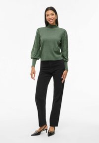 Maglione verde con collo alto e maniche voluminose, abbinato a jeans neri a gamba dritta e décolleté neri a punta.