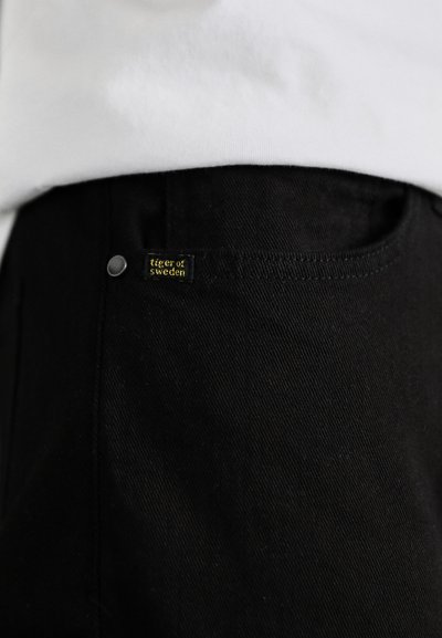 Pantalones de mezclilla negra con una textura suave, que presentan una pequeña etiqueta cosida en amarillo que dice "tiger of sweden" y un botón metálico.