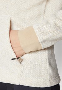 Light-colored, textured sweater with a contrasting tan cuff, features a side pocket with a drawstring. Subtle pattern throughout the fabric.