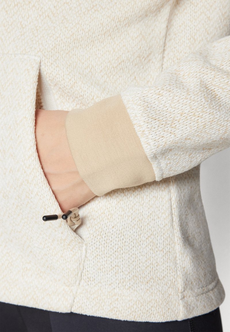 Pull léger de couleur claire et texturé avec un poignet en tan contrasté, équipé d'une poche latérale avec un cordon de serrage. Motif subtil sur l'ensemble du tissu.