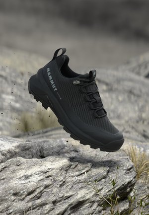 Czarny but turystyczny Mammut z wytrzymałą podeszwą unoszący się nad skalistym terenem z rzadką trawą i rozmytym naturalnym tłem.