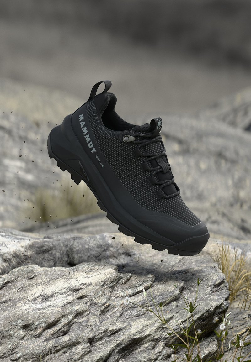 Pantof de drumeție Mammut negru cu talpă robustă, plutind deasupra unui teren stâncos cu iarbă rară și fundal natural estompat.