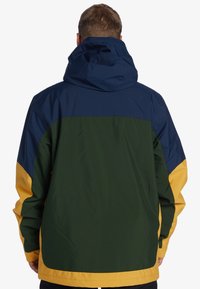 Chaqueta resistente al agua con un diseño en bloques de color en azul marino, verde y amarillo brillante, con capucha y puños elásticos.