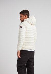 Giacca piumino bianca con cappuccio, caratterizzata da cuciture orizzontali e una toppa con il logo sul braccio sinistro. Abbinata a pantaloni neri.