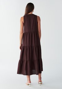 Mørkebrun, ærmeløs maxikjole med en vid, lagdelt design og en lige ryg. Fremstillet af let stof med en subtil tekstur.