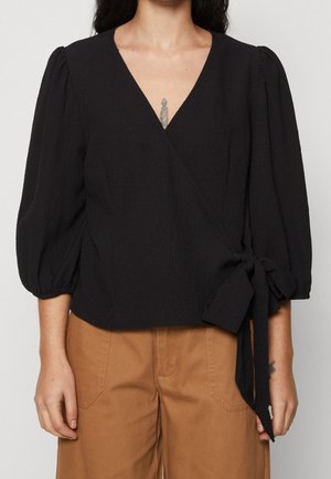 Femme portant un chemisier noir enveloppant avec des manches bouffantes noué sur le côté, et un pantalon marron taille haute.