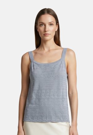 Donna che indossa un top lavorato a maglia smanicato grigio chiaro con sottili spalline e sottili motivi testurizzati, in piedi su uno sfondo bianco uniforme.