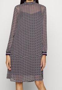 Langärmliges, durchscheinendes Kleid mit einem schwarz-lila geometrischen Muster, das einen hohen Kragen und gestreifte Ärmelabschlüsse in Schwarz, Lila und Pink aufweist.