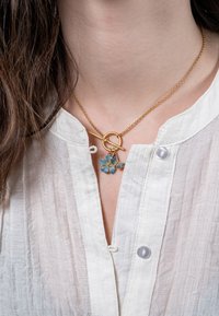 Kuldne kaelakee ümarate kinnitusplokkidega, millel on sinised lillemotiivid. Ahel on õrn, täiustades valget tekstuuriga pluusi.