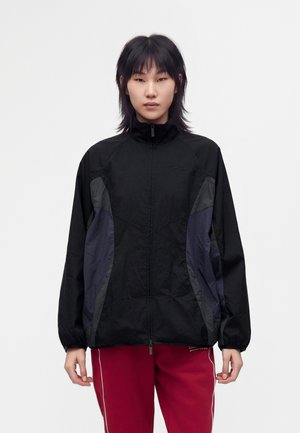 Personne aux cheveux noirs mi-longs porte une veste noire à fermeture éclair avec des panneaux latéraux gris et un pantalon rouge avec des bandes blanches sur les côtés.