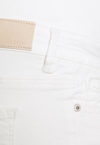 Pantalones de mezclilla blanca con una etiqueta de cuero marrón claro con la palabra "ESPRIT" grabada. Incluyen herrajes plateados y detalles de costura visibles.