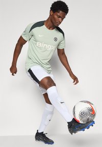 Un joueur de football masculin portant un maillot d'entraînement Chelsea vert clair contrôle le ballon en l'air du pied droit, vêtu de shorts blancs et de crampons.