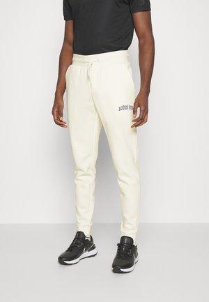 Mężczyzna w kremowych spodniach dresowych z logo "BJÖRN BORG" na udzie, czarnej koszulce i czarnych sneakersach Nike stoi na szarej podłodze.