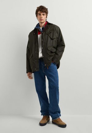 Joven con chaqueta verde oscuro, sobre una camisa blanca y roja, con vaqueros azules y zapatillas marrones, de pie frente a un fondo claro y liso.