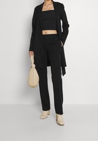Un costume noir comprenant un manteau long ajusté et un pantalon fitted, associé à un crop top noir. Complété par des bottes à talons beiges et un sac.