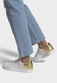 Vita sneakers med gyllene detaljer, med ett perforerat mönster. Sulan är vit och materialet verkar slätt och stilrent.