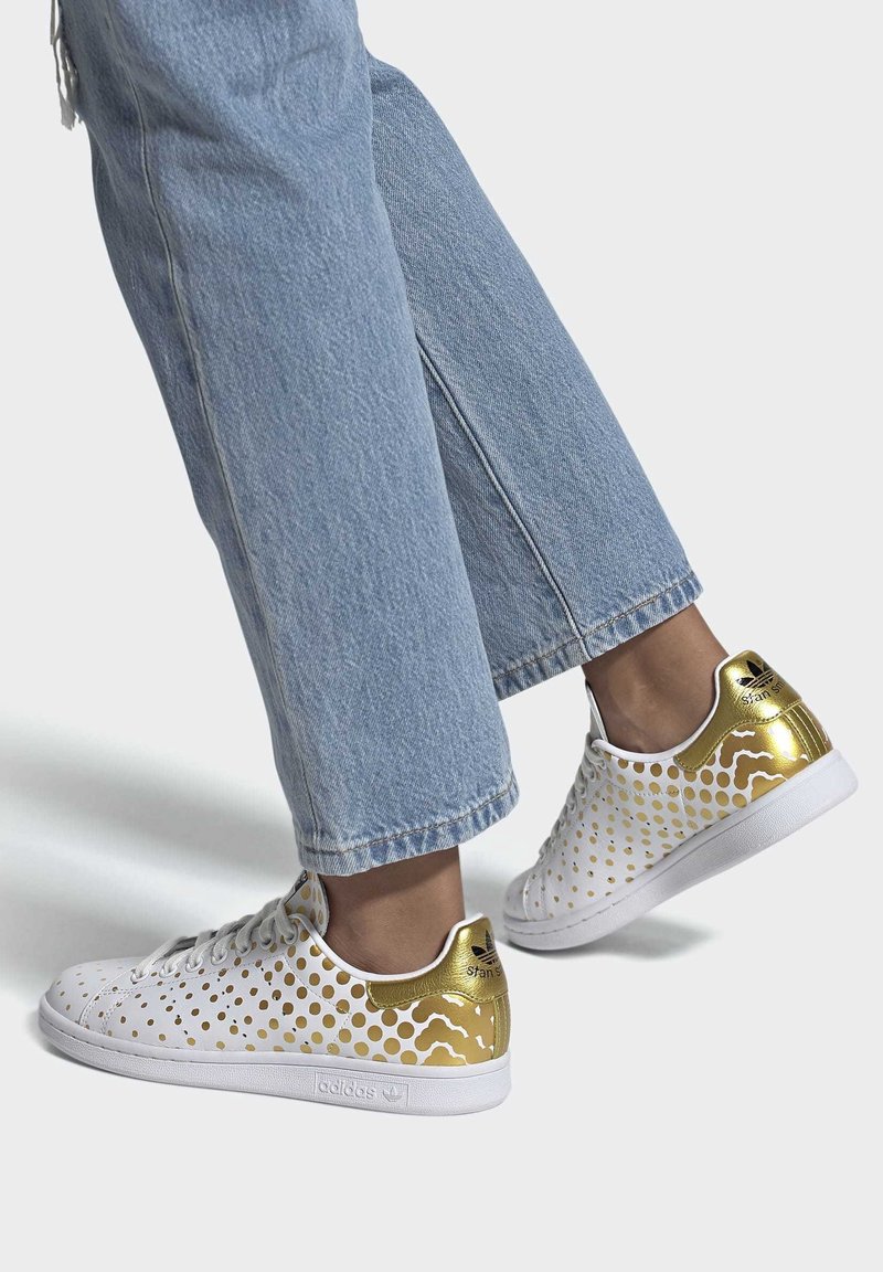 Vita sneakers med gyllene detaljer, med ett perforerat mönster. Sulan är vit och materialet verkar slätt och stilrent.