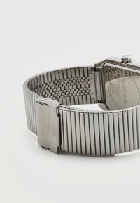 Ασημένια μεταλλική μπάντα ρολογιού με κούμπωμα που φέρει το λογότυπο κροκοδείλου, προσαρμοσμένη σε ορθογώνια θήκη ρολογιού που δείχνει λεπτομέρειες μοντέλου και αντοχής στο νερό.