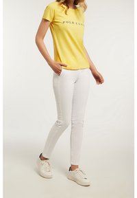 Żółta bawełniana koszulka z napisem "POLO CLUB", zestawiona z białymi jeansami slim-fit i białymi trampkami. Prosty, codzienny strój o czystym wzornictwie.