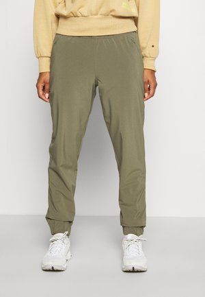 Personne portant un pantalon de jogging vert olive avec des poignets élastiques et des baskets blanches, debout devant un fond clair uni.
