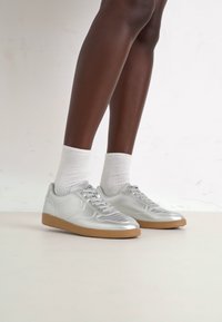Silberne Sneakers mit glatter Oberfläche, Gummisohle und strukturierten Akzenten. Kombiniert mit weißen Socken, die ein sportliches Design zeigen.