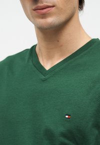 Κοντινό πλάνο ενός άντρα που φοράει πράσινο μπλουζάκι με V-λαιμόκοψη και ένα μικρό ορθογώνιο λογότυπο στην αριστερή πλευρά του στήθους.