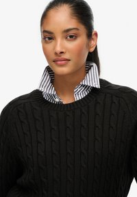 Zwarte kabelgebreide trui met een geribde halslijn, gedragen over de kraag van een wit met marineblauw gestreept overhemd. Elegante textuur en een aansluitende pasvorm.